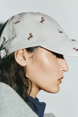 Gorra de sarga confeccionada en algodón, con visera curva y ajuste posterior. Presenta un diseño de color gris con múltiples bordados pequeños de perros salchicha en diversos colores.