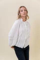 Camisa blanca de tela liviana con detalles de encaje y terminaciones caladas. Presenta cuello con leve volado, botonera frontal y mangas largas abullonadas con puños ajustados.