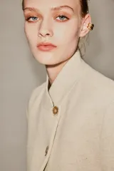 Blazer de corte estructurado en color beige, con cuello mao y cierre frontal cruzado mediante botones. Presenta mangas largas con detalle de botones en los puños y bolsillos de vivo en el frente.