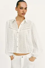 Blusa corta de color blanco con diseño de bordados perforados, cuello solapa, cierre frontal con botones y mangas largas abullonadas.