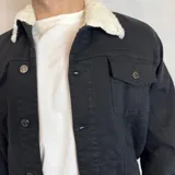 Chaqueta de jean negra con cuello de corderito blanco, cierre frontal con botones metálicos, bolsillos con solapa en el pecho y bolsillos laterales.