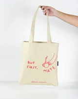 Bolso de mano tipo tote bag color crudo, con asas largas y estampado de una mano sirviendo mate en color rojo.