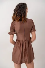 Vestido corto color marrón chocolate, con cuerpo fruncido tipo nido de abejas, cuello alto con volados y mangas cortas abuchonadas con terminación en volado. Presenta falda evasé con vuelo.