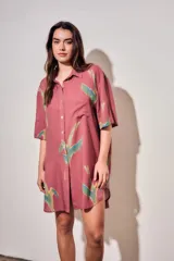 Vestido camisero corto de color rosado con estampado de aves abstractas en tonos verdes y azules. Cuenta con cuello camisero, cierre frontal con botones, mangas cortas y un bolsillo aplicado en el pecho.