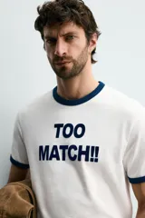 Camiseta de corte regular con cuello redondo y mangas cortas con ribetes en contraste de color. Presenta una estampa frontal con la frase 'TOO MATCH!!' en tipografía destacada.