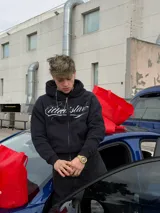 Campera con capucha negra, con cierre frontal completo y bolsillos estilo canguro. Presenta un estampado gráfico en blanco con letras cursivas en el pecho y diseño holgado.