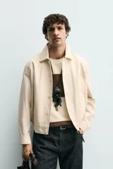 Cazadora boxy fit color beige, confeccionada en tejido de algodón y lino. Presenta cuello solapa, manga larga con puño abotonado, bolsillos de vivo en la cadera y bajo con elásticos laterales. Cierre frontal de botonadura.