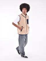 Chaleco de polar tipo corderito en color beige, con cuello alto y cierre frontal completo. Presenta un bolsillo de parche en el pecho en color azul marino con cierre de cremallera y ribetes en contraste en el mismo tono en sisas, cuello y bolsillos laterales.