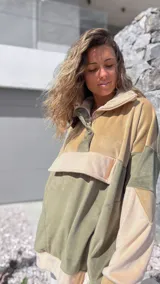 Buzo tipo canguro de diseño color block en tonos verde oliva y beige, confeccionado en tejido de textura aterciopelada. Presenta cuello alto con cierre frontal parcial, capucha y un bolsillo frontal grande con solapa.