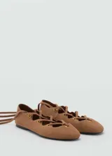 Zapatos chatitos de piel de serraje color marrón, con diseño de tiras cruzadas en el empeine, ojales metálicos dorados y cintas largas para atar alrededor del tobillo. Presentan punta redonda y suela plana.