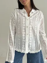 Camisa blanca de manga larga con delicados bordados florales verticales y detalles de encaje. Presenta cuello clásico, cierre frontal con botones y terminaciones festoneadas en los puños y el ruedo.