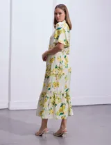 Vestido largo camisero con estampado floral amarillo y verde sobre fondo blanco. Tiene cuello camisero, mangas cortas con volados, cintura con lazo ajustable y falda escalonada con volados.