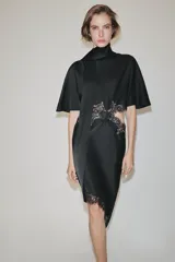 Vestido midi de satén negro con cuello subido, manga corta tipo capa y pañuelo incorporado en el mismo tejido. Presenta aberturas laterales decoradas con encaje a tono y bajo asimétrico con detalles de encaje. Cierre en la espalda con abertura y botones forrados.