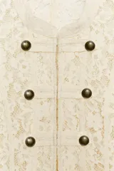 Chaqueta corta de encaje color crudo, con cuello mao y cierre frontal de ganchos metálicos decorados con botones estilo militar. Presenta mangas largas con detalles de botones en los puños.