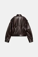 Cazadora corta de efecto piel color marrón oscuro, con cuello subido y cierre frontal oculto con cremallera y botones a presión. Presenta mangas largas con puños y pliegues, trabillas en los hombros, bolsillos laterales y bajo abullonado. El tejido tiene un efecto lavado.