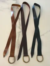 Cinto de cuero flexible y envolvente con diseño minimalista, que se anuda mediante una hebilla metálica en forma de D. El extremo presenta el logo de la marca grabado.