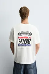 Camiseta blanca de corte boxy fit, cuello redondo y manga corta. Presenta un parche bordado con las letras "NY" y un diseño abstracto en negro en el pecho.