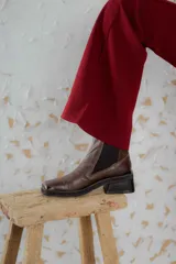 Botas tipo chelsea de cuero color marrón oscuro con acabado brillante. Presentan punta cuadrada con detalle de puntera metálica, elásticos laterales y taco cuadrado bajo.