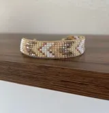 Pulsera artesanal tejida en telar con cuentas Miyuki en tonos beige, blanco, gris y dorado, formando un diseño geométrico. Cuenta con cierre regulable.