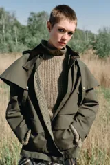 Trench de estilo encerado color verde militar con cuello de solapa en pana marrón, capucha extraíble y capa superior. Presenta cierre frontal con cremallera y botones a presión ocultos, bolsillos delanteros de plastrón con solapa y mangas largas con trabillas.