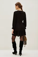 Vestido corto color negro, con escote en V decorado con volados, mangas largas con puños elásticos y falda con volado en el ruedo. Incluye un lazo para ajustar en la cintura.