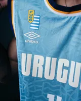 Camiseta de básquet celeste con cuello y sisas en azul marino. Estampado frontal con la palabra "Uruguay" en blanco y el logo de Antel. Incluye logo de Umbro y el escudo de Uruguay. No incluye número ni parche.