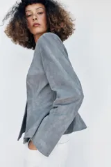 Chaqueta de cuero gamuzado color gris, con cuello redondo, manga larga, cierre frontal con botones oscuros y bajo con detalle peplum.