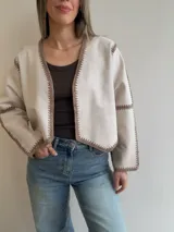 Chaqueta de efecto gamuza color beige, con diseño abierto sin cierres, mangas largas y terminaciones artesanales bordadas en tono marrón en los bordes y costuras.