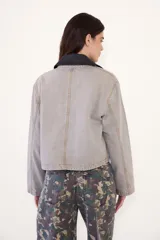 Campera corta de jean color visón, con cuello desmontable de tela engomada, cierre frontal y bolsillos plaqué con solapa en el pecho. Cuenta con un parche bordado en el delantero.