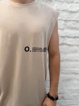 Musculosa de color beige con cuello redondo y corte holgado. Presenta un estampado minimalista en negro en el lateral del pecho con el nombre de la marca Osten Studio y coordenadas geográficas.