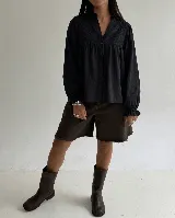 Blusa negra de manga larga con cuello mao, cierre frontal de botones y detalle de bordado geométrico en la parte superior delantera. Presenta un corte holgado con puños elásticos con volado.