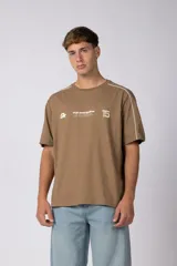 Remera de algodón color marrón con cuello redondo y mangas cortas. Presenta un diseño gráfico estampado en el pecho con texto y números, además de líneas blancas decorativas que recorren los hombros y mangas.
