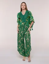 Blusa verde de gasa con transparencia y estampado metalizado dorado, de silueta holgada y mangas cortas. Presenta escote en V con pechera tejida en crochet y apliques de borlas colgantes.