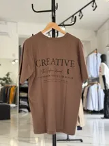 Remera oversize color beige, con diseño estampado en la espalda que incluye tipografía grande y texto descriptivo en negro.