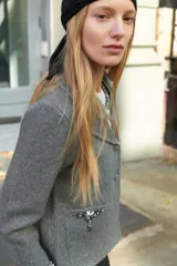 Chaqueta de punto gris con cuello solapa, manga larga y cierre frontal con botones forrados. Presenta bolsillos delanteros de plastrón y detalles de abalorios joya en los bolsillos y en los puños.