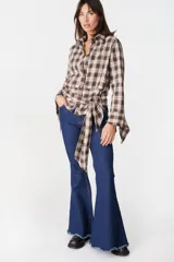 Camisa de corte holgado con estampado de cuadros en tonos marrones y beige. Presenta cuello clásico, cierre frontal con botones y mangas largas con aberturas laterales que permiten anudar la prenda en la cintura.