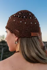Casquete tejido a mano en crochet color marrón con acabado brillante, decorado con perlas blancas distribuidas por toda la superficie. Incluye una etiqueta de cuero con la marca Soriame.