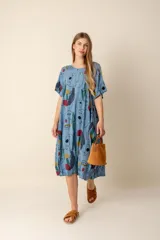 Vestido largo estampado con flores en tonos violeta, naranja, verde y azul sobre un fondo beige claro. Tiene cuello en V y mangas cortas.