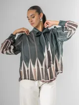 Camisa de manga larga con cuello alto y lazo para atar, confeccionada en tela liviana con estampado geométrico en tonos verde oscuro, blanco, beige y marrón.