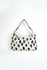 Cartera estilo hobo de color crudo con estampado de manchas negras abstractas, cierre superior y correa de hombro ajustable de cuero negro.