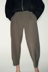 Pantalón de tiro medio con pinzas delanteras, bolsillos laterales y falsos bolsillos de vivo en la parte trasera. Presenta un corte holgado que se estrecha hacia el tobillo, con bajo acabado en pliegues y cierre frontal con cremallera, botón interior y gancho.