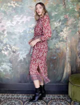 Vestido camisero midi de gasa estampada con flores rojas y blancas sobre fondo marrón. Tiene cuello con solapa, abertura central con botones, mangas largas y lazo de ajuste en la cintura.