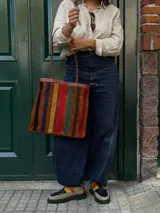 Cartera tipo tote de cuero con diseño de franjas verticales multicolores en tonos tierra, rojo y azul. Cuenta con correas trenzadas de cuero y cierre superior.