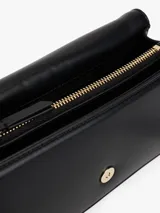 Cartera tipo bandolera de color negro, con acabado liso y solapa frontal. Presenta herrajes metálicos dorados, incluyendo un cierre con el monograma TH y un logo de Tommy Hilfiger en bajo relieve.