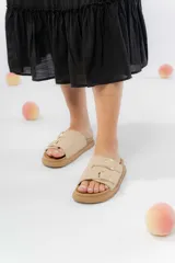 Sandalias color beige con dos tiras anchas con hebillas metálicas doradas, confeccionadas en material gamuzado sintético. Suela acolchada y antideslizante.