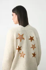 Chaqueta de punto color crema, con diseño abierto, mangas largas y flecos en el ruedo. Presenta un aplique bordado en forma de estrella en uno de los bolsillos frontales.
