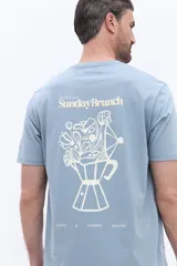 Remera de manga corta color azul grisáceo con cuello redondo y calce regular. Presenta un estampado gráfico en la espalda que combina una cafetera italiana con flores y el texto Sunday Brunch.