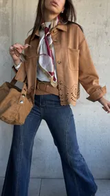 Chaqueta de gamuza color camel con cierre frontal de botones, cuello clásico y detalle de calados decorativos en el ruedo. Presenta bolsillos frontales y mangas largas con puños.