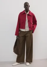 Chaqueta corta de algodón en color rojo, con cuello camisero, manga larga y cierre frontal de botones oscuros. Presenta dos bolsillos con solapa en el frente.