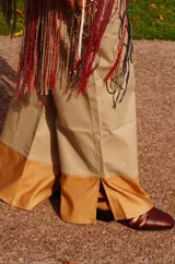 Pantalón ancho de tiro alto color beige, con pliegues frontales y botones visibles en la cintura. El ruedo del pantalón tiene un detalle de terminación en cuero color caramelo.